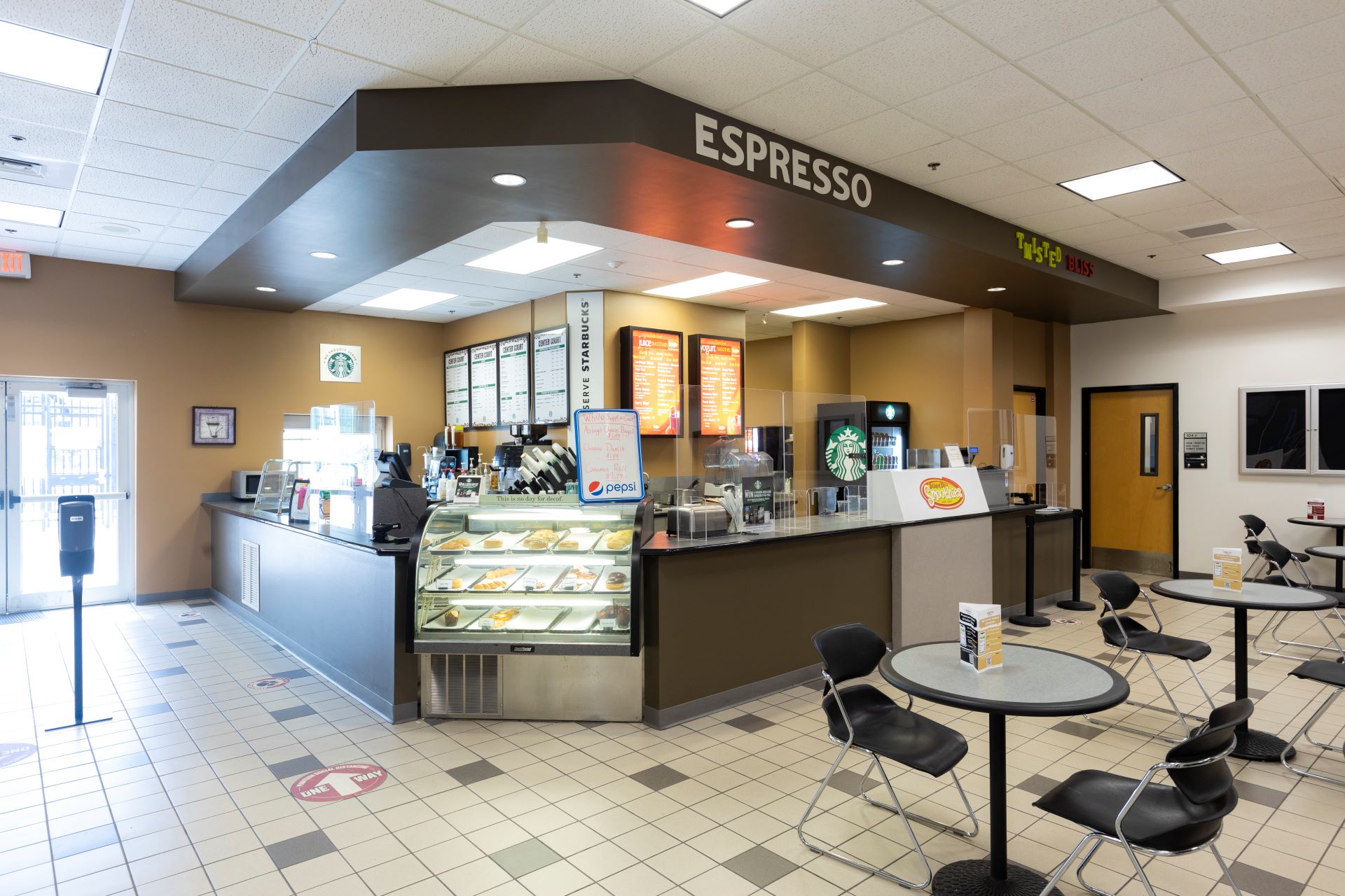 An interior view of a cafe area at Florida Tech, featuring an espresso counter with a Starbucks menu, display case with pastries, and seating area with tables and chairs. The cafe also includes signage for Twisted Bliss and an entrance door to the left